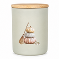 A cylindrical ceramic jar with a light sage green body, a natural wood lid, and a printed illustration of stacked pumpkins and a small broom on the front.