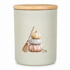 A cylindrical ceramic jar with a light sage green body, a natural wood lid, and a printed illustration of stacked pumpkins and a small broom on the front.