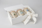 Three cream-colored soy candles with light wood lids are lined up on a white marble countertop. The candles feature watercolor illustrations and labels: 'Memories Christmas Night' with a Christmas tree, 'Memories Summer Drive-In' with a blue van and palm trees, and 'Memories Mental Coconut' with a bluebird.