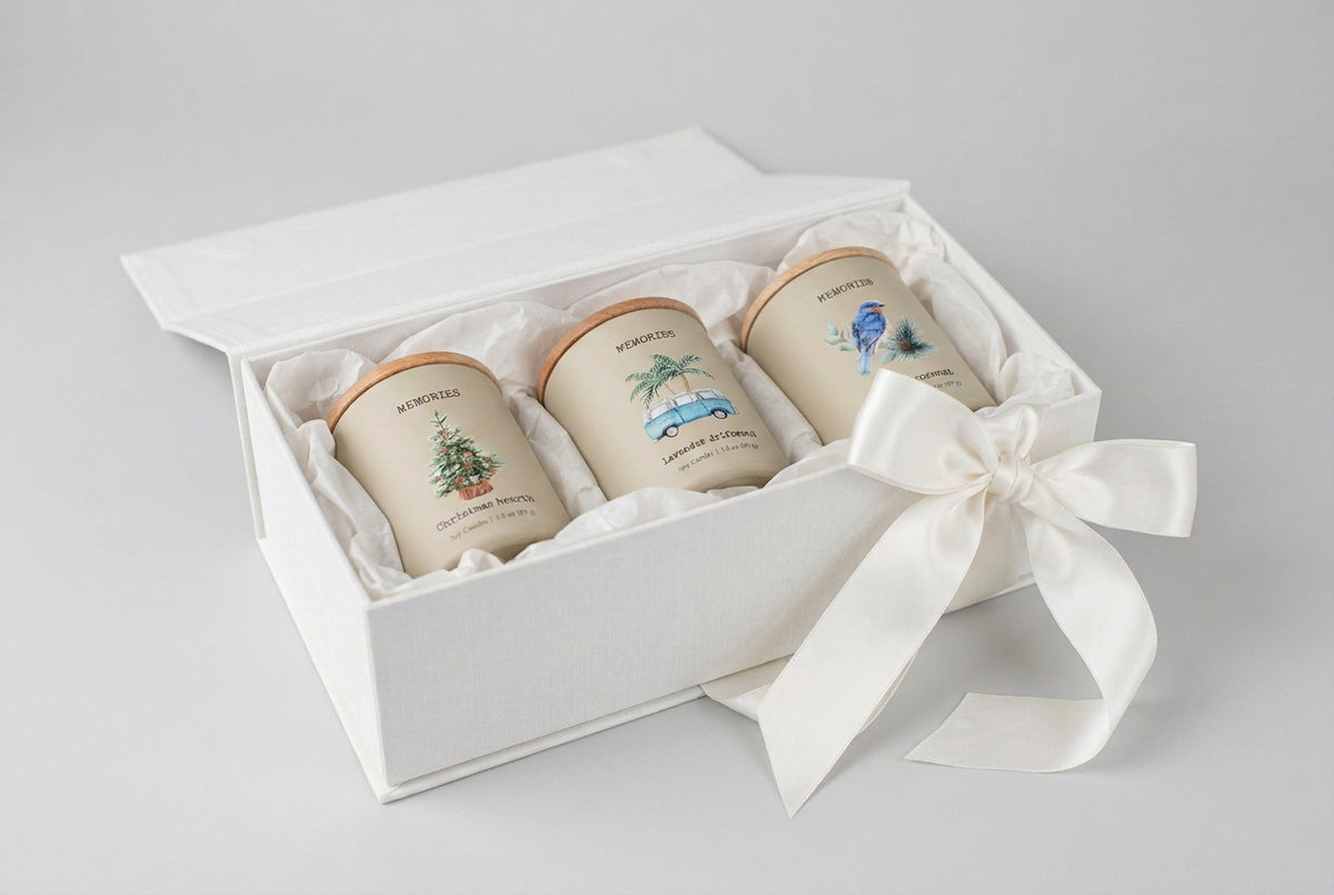 Three cream-colored soy candles with light wood lids are lined up on a white marble countertop. The candles feature watercolor illustrations and labels: 'Memories Christmas Night' with a Christmas tree, 'Memories Summer Drive-In' with a blue van and palm trees, and 'Memories Mental Coconut' with a bluebird.