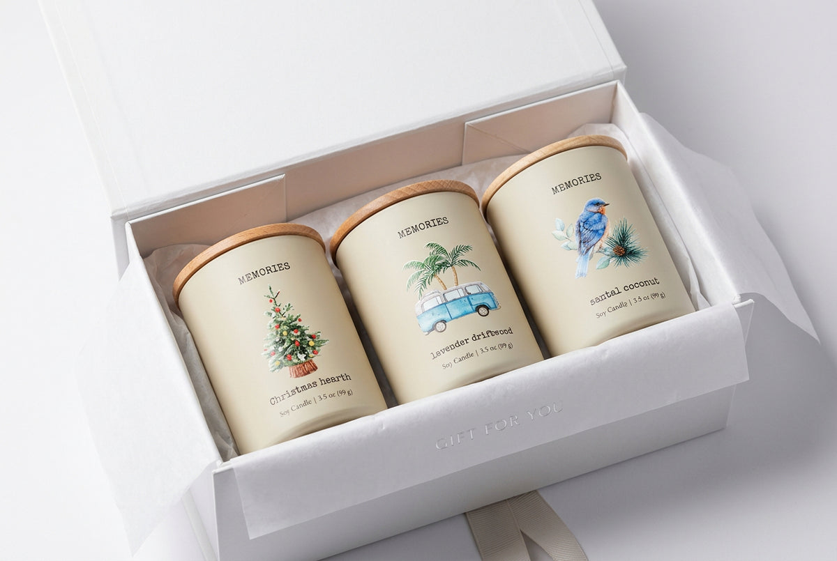 Three cream-colored soy candles with light wood lids are lined up on a white marble countertop. The candles feature watercolor illustrations and labels: 'Memories Christmas Night' with a Christmas tree, 'Memories Summer Drive-In' with a blue van and palm trees, and 'Memories Mental Coconut' with a bluebird.